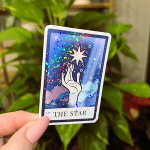 May include: A tarot card with a blue, purple and white background. A hand reaches up to touch a white star. The card reads "THE STAR".