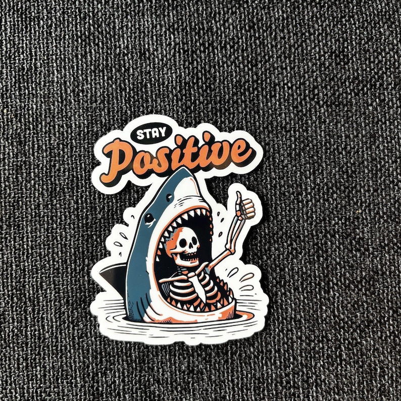 May include: A sticker featuring a cartoon shark with a skeleton inside, giving a thumbs-up. The shark is blue and white, with an open mouth. The words "Stay Positive" are above the shark in orange and white.