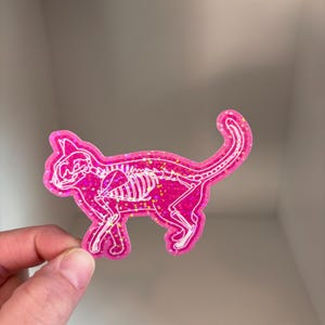 May include: Pink glitter sticker of a cat skeleton. The sticker is white and pink with a holographic finish.