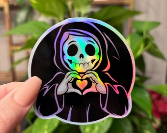 May include: A holographic sticker featuring a cartoon skeleton wearing a black hooded robe and making a heart shape with its hands. The skeleton has a rainbow iridescent effect.