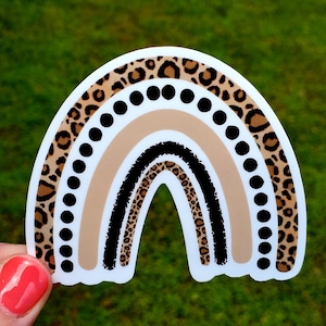 May include: A rainbow sticker with a brown and black leopard print design. The rainbow has a black outline and is decorated with black dots.
