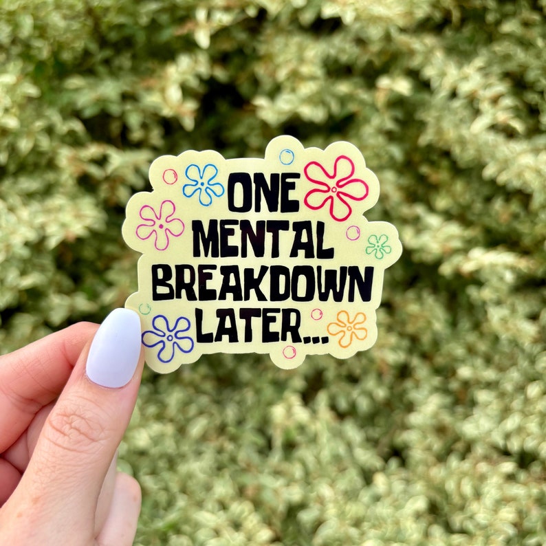 May include: A yellow sticker with the text "ONE MENTAL BREAKDOWN LATER..." in black and red. The sticker is decorated with colorful flower and bubble designs. The background is a blurred green.