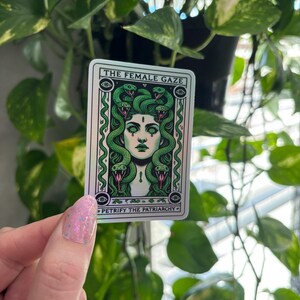 May include: A sticker with a green and white illustration of a woman with snakes for hair. The text on the sticker reads "The Female Gaze" and "Petrify the Patriarchy".