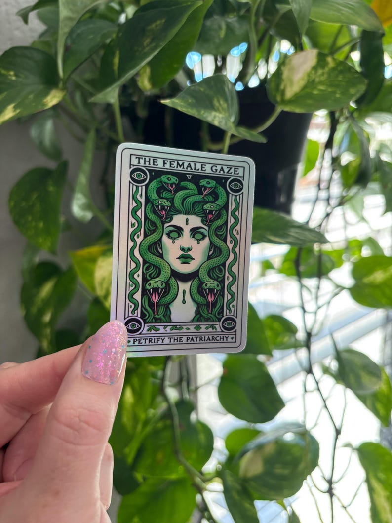 May include: A sticker with a green and white illustration of a woman with snakes for hair. The text on the sticker reads "The Female Gaze" and "Petrify the Patriarchy".