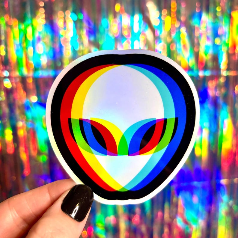 May include: A holographic sticker featuring an alien head design with a 3D effect. The alien's eyes and head are outlined in red, yellow, and blue, with a black border. The sticker is held against a shimmering, rainbow-colored background.