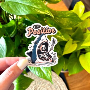 May include: A sticker with a cartoon shark design. The shark's mouth is open, revealing a skeleton giving a thumbs-up. The sticker has the words "Stay Positive" in a cursive font.