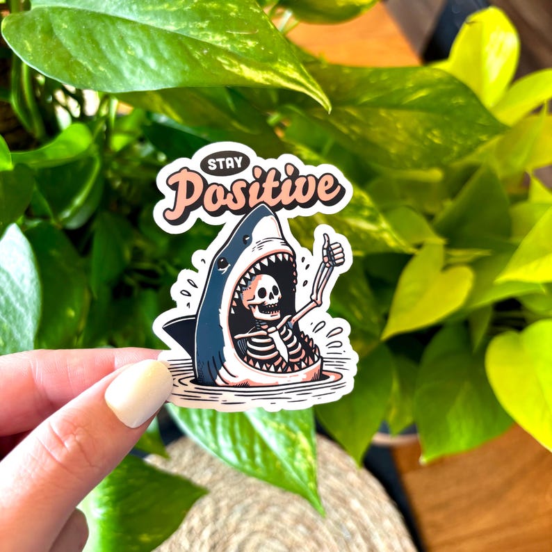 May include: A sticker with a cartoon shark design. The shark's mouth is open, revealing a skeleton giving a thumbs-up. The sticker has the words "Stay Positive" in a cursive font.