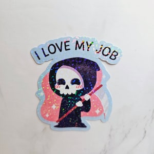 Op de afbeelding: Een holografische sticker met een cartoon Grim Reaper met een zeis, die een zwarte kapmantel en een roze halo draagt. De tekst "I LOVE MY JOB" staat op een blauwe banner boven de Grim Reaper.