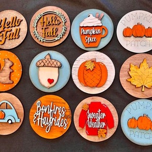 May include: Twelve round wooden signs with various fall-themed designs. The signs feature images of pumpkins, acorns, squirrels, a leaf, a car, a sweater, and text that says "Hello Fall", "Pumpkin Spice", "Pumpkin Patch", "Bonfires & Hayrides", and "Sweater Weather".