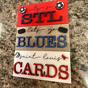 May include: A wooden sign with three tiers, each featuring a different St. Louis sports team. The top tier is red and says "city sc STL" with a soccer ball. The middle tier is brown and says "lets go BLUES" with a hockey puck. The bottom tier is white and says "saint louis CARDS" with a baseball and a cardinal.