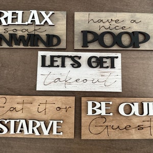 May include: Five wooden signs with different sayings. The signs are all different colors and have a rustic look. The sayings on the signs are: "Relax soak unwind", "Have a nice poop", "Let's get takeout", "Eat it or starve", and "Be our guest".