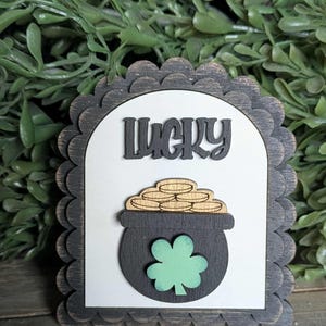 May include: Wooden St. Patrick's Day decoration with the word "Lucky" in black script. The design features a pot of gold, a green shamrock, and a scalloped border. The background is white.