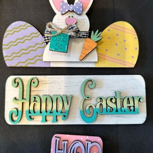 May include: Wooden Easter decorations featuring a bunny with a bow, carrot, and eggs. The bunny has a price tag that says "EGGS 5¢". Additional signs read "Happy Easter" and "HOP".