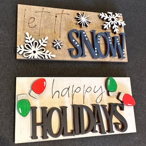May include: Two wooden signs with a winter holiday theme. The top sign is brown with white snowflakes and the text "let it snow". The bottom sign is white with green and red light bulbs and the text "happy holidays".