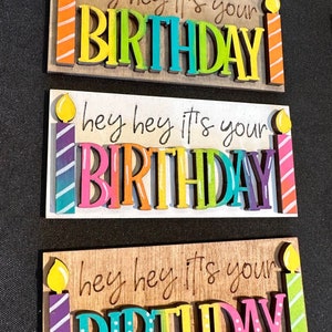 May include: Three wooden signs with the text "hey hey it's your BIRTHDAY" in colorful letters and a candle on each side. The candles are yellow with orange, purple, and green stripes.