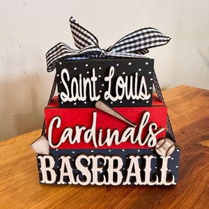 May include: A three-tiered wooden block set with the words "Saint Louis Cardinals Baseball" in white lettering. The blocks are painted black, red, and blue with white polka dots. A black and white checkered ribbon is tied around the top block.