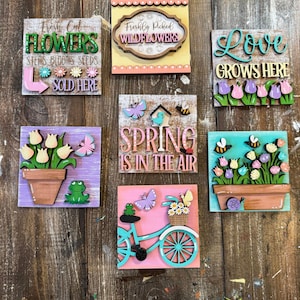 May include: Six wooden signs with spring-themed designs. The signs feature flowers, butterflies, bees, a birdhouse, a bicycle, and the words "Fresh Cut Flowers", "Freshly Picked Wildflowers", "Love Grows Here", "Spring is in the Air", and "Sold Here".