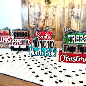 May include: Four tiered wooden Christmas ornaments with festive holiday themes. The ornaments are decorated with red, green, white, and black paint and feature the words "Cookie Icing Gingerbread", "Cocoa Peppermint Marshmallow", "Santa Ho Ho Ho North Pole", and "Trees Farm Fresh Christmas".