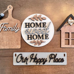 May include: Four wooden signs with black and gray lettering. The signs read "Family", "Home Sweet Home", "Our Happy Place", and a house with a heart.