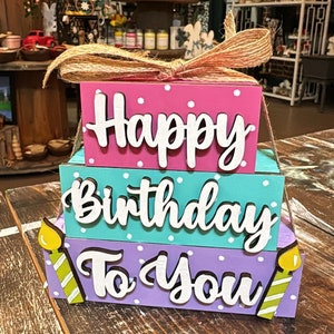 May include: A three-tiered wooden birthday sign with the words "Happy Birthday To You" in white letters on a pink, teal, and purple background with white polka dots. The sign is decorated with two yellow candles with green and white stripes.