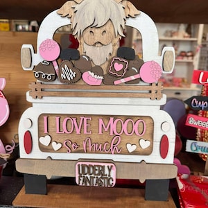 May include: A wooden cutout of a Highland cow in a pickup truck with a pink and white "I Love Moo So Much" sign and a pink and white "Udderly Fantastic" license plate. The truck bed is filled with chocolate hearts, candies, and lollipops.