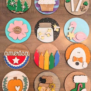 May include: A collection of wooden round ornaments with various designs. Includes a cupcake, a flamingo, a pumpkin, a scarecrow, and a Christmas stocking. Other designs include a flower, a jar with bees, and a patriotic "America" design.