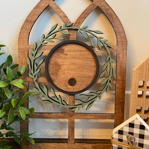 May include: A wooden window frame with a green wreath and a round cutout in the center. The window frame is brown and has a distressed look. The wreath is made of green leaves and has a black outline. The cutout is round and has a brown wood grain.
