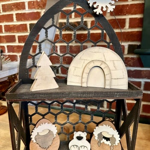 May include: A two-tiered wooden shelf with a black and white honeycomb pattern. The top shelf has a white wooden igloo, a white wooden Christmas tree, and a silver snowflake. The bottom shelf has two brown wooden figures wearing white fur hats and a white wooden polar bear.