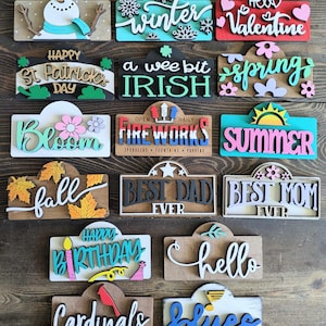 May include: A collection of 15 wooden signs with various holiday and seasonal themes. The signs are painted in bright colors and feature festive designs, such as snowflakes, hearts, shamrocks, and fireworks. The signs are perfect for decorating a home or business for any occasion.