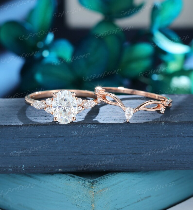 May include: A rose gold engagement ring set with an oval-shaped center stone and smaller pear-shaped stones on the band. The matching wedding band features a delicate, vine-like design with pear-shaped stones.