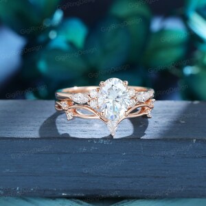 May include: A rose gold engagement ring set with a pear-shaped center stone and smaller round stones on the band. The ring is set on a dark wooden surface.