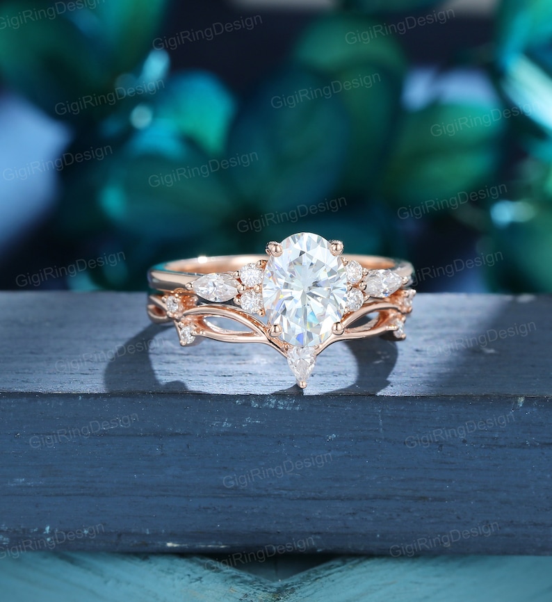 May include: A rose gold engagement ring set with a pear-shaped center stone and smaller round stones on the band. The ring is set on a dark wooden surface.