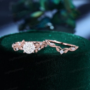 Könnte beinhalten: Ein Verlobungsring-Set aus Roségold mit einem runden Brillantschliff-Mittelstein, flankiert von blattförmigen Metallakzenten und kleinen runden Steinen, kombiniert mit einem passenden geschwungenen Ehering, der mit zarten Blättern und kleinen Steinen verziert ist.