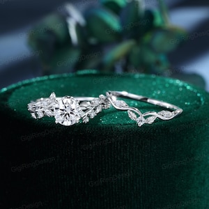 May include: A silver-toned engagement ring set featuring a round brilliant-cut center stone flanked by leaf-shaped accents and a matching curved wedding band with small round stones and leaf details, both crafted with a polished metallic finish.
