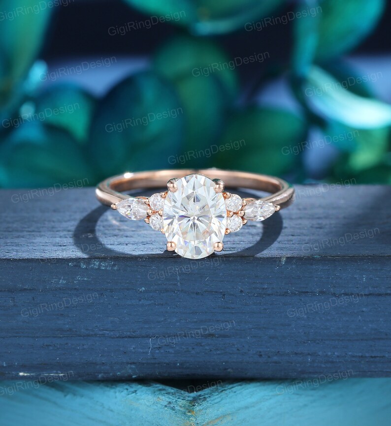 May include: A rose gold engagement ring with an oval-shaped center stone and pear-shaped accent stones. The ring is set on a white background.