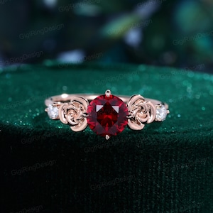 May include: Rose gold ring featuring a large, round, deep red gemstone as the centerpiece. The ring is adorned with rose-shaped accents on either side of the central stone, and small, clear accent stones. The ring is set against a dark green background.