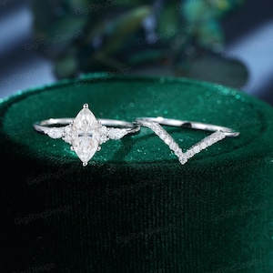 May include: A silver-toned ring set featuring a central marquise-cut stone flanked by two smaller pear-shaped stones on each side, paired with a matching V-shaped chevron band encrusted with a row of small round stones along the top edge.