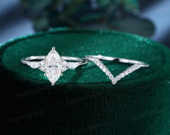 Op de afbeelding: Een zilverkleurige ringenset met een centrale steen in marquise-slijpvorm, geflankeerd door twee kleinere peervormige stenen aan weerszijden, gecombineerd met een bijpassende V-vormige chevronring bezet met een rij kleine ronde steentjes.