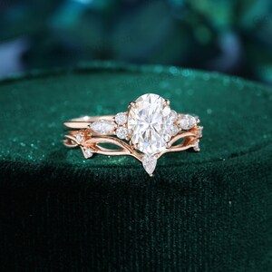 May include: A rose gold engagement ring set with a large oval-shaped center stone and smaller pear-shaped accent stones. The ring band features a delicate vine design with small leaves.