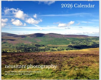 May include: A 2026 calendar featuring a scenic landscape with rolling hills, green valleys, and a vibrant blue sky dotted with clouds. The foreground shows a field of purple and brown vegetation. The text "2026 Calendar" is at the top, and "neusitzer photography" is at the bottom.