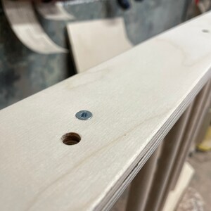 May include: Close-up of a light wood shelf with a screw and a hole drilled into it. The shelf is part of a larger piece of furniture.