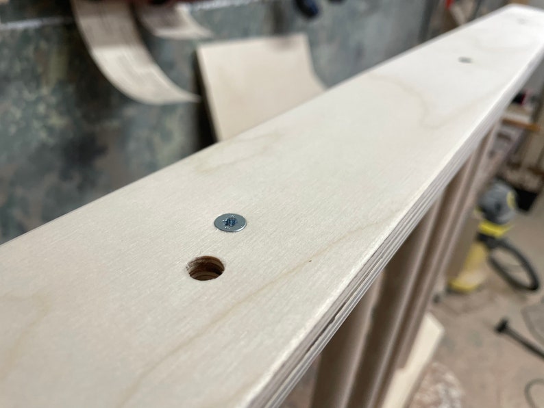 May include: Close-up of a light wood shelf with a screw and a hole drilled into it. The shelf is part of a larger piece of furniture.