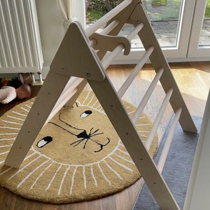 May include: A wooden triangular climbing frame with a ladder and a rope hanging from the top. The frame is standing on a beige rug with a lion face design.