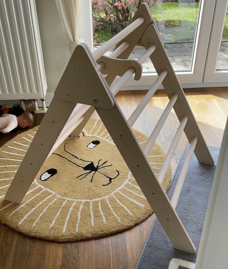 May include: A wooden triangular climbing frame with a ladder and a rope hanging from the top. The frame is standing on a beige rug with a lion face design.