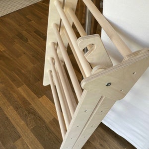 May include: A wooden climbing ladder for children, made of natural wood with rounded edges. The ladder is folded and leaning against a white surface.