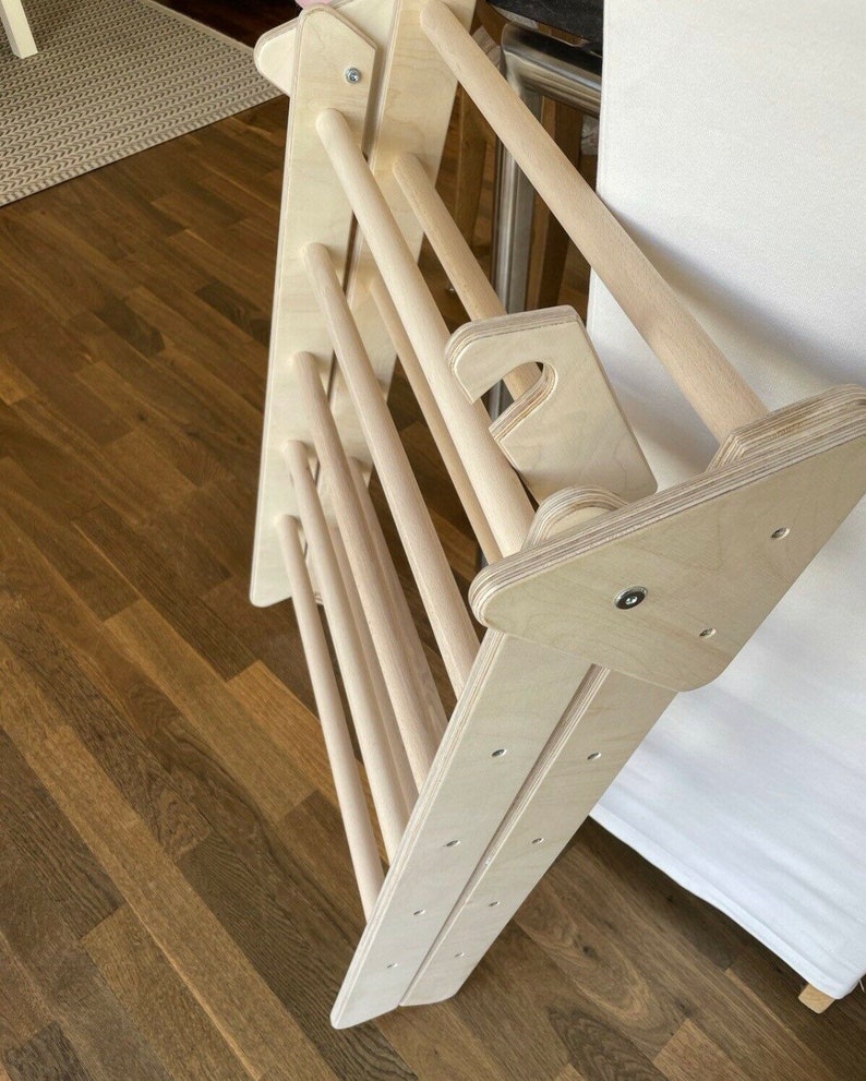 May include: A wooden climbing ladder for children, made of natural wood with rounded edges. The ladder is folded and leaning against a white surface.