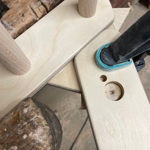 May include: A close-up of a wooden frame being assembled with a clamp. The frame is made of light wood and has a hole drilled in it. A hammer is visible in the foreground.