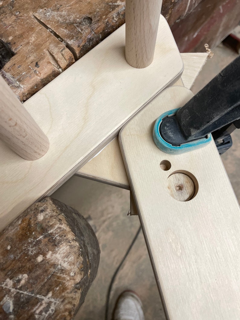 May include: A close-up of a wooden frame being assembled with a clamp. The frame is made of light wood and has a hole drilled in it. A hammer is visible in the foreground.