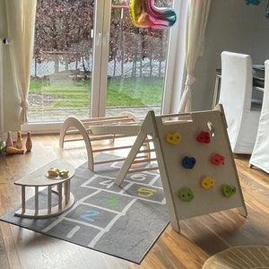 May include: A wooden play structure with a climbing wall and a slide. The structure is set up in a room with a gray rug with a hopscotch design. There are colorful balloons in the background, including a large rainbow-colored number two balloon.