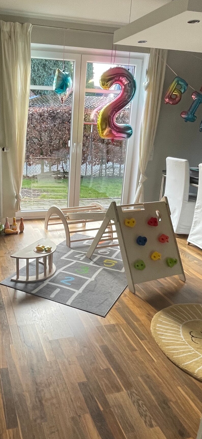May include: A wooden play structure with a climbing wall and a slide. The structure is set up in a room with a gray rug with a hopscotch design. There are colorful balloons in the background, including a large rainbow-colored number two balloon.